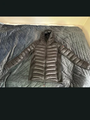 Svart dunjacka från Canada Snow - Snygg svart dunjacka från Canada Snow med huva och quiltad design. Jackan har dragkedja framtill, två fickor med dragkedja och en patch med logga på ärmen. Perfekt för kalla vinterdagar och riktigt skön att bära.