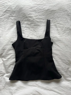 Svart topp från Zara med rosett i ryggen - Stilren svart linnetopp med breda axelband och snygg rosettdetalj i ryggen. Toppen har figurnära passform och lätt utsvängd nederkant, perfekt för en trendig look. Materialet känns mjukt och följsamt mot huden.