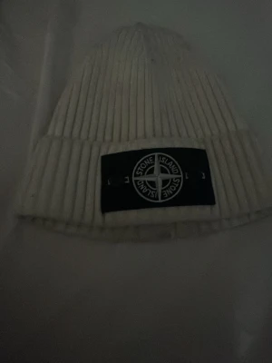 Vit ribbstickad mössa Stone Island - Snygg vit ribbstickad mössa från Stone Island med klassisk svart logotyp-patch framtill. Mössan har uppvikt kant och är tillverkad i ett mjukt och värmande material som passar perfekt för kyliga dagar.
