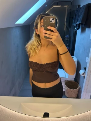Brun bandeau topp med spetsdetalj - Snygg brun bandeau topp med spets upptill. Toppens material är stretchigt och sitter tight mot kroppen. Perfekt för dig som gillar en enkel men ändå trendig look. Ärmlös och axelbandslös modell.