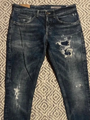 Blå slitna jeans från dondup - Säljer ett par blå jeans från dondup med slitningar och hål på ena benet. Jeansen har fem fickor, smal passform och klassisk knappgylf. Materialet är denim med coolt distressed look och detaljerade sömmar. De är som nya skick 9/10 nypris ligger på 3600