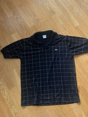Svart rutmönstrad piké från Lacoste  - Snygg svart pikétröja från Lacoste med vitt/orange rutmönster. Jag är 182 lång och den sitter baggy och lite långt. Möts i Stockholm 