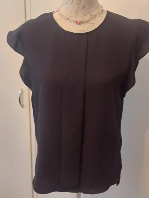 Svart blus med volangärm - Snygg svart blus med rund halsringning och volangärmar. Blusen har en rak passform och ett diskret veck framtill som ger en stilren look. 
