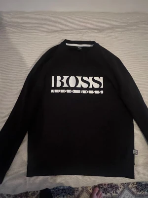 Hugo boss  - Svart sweatshirt från Hugo Boss med stor vit logga på bröstet. Tröjan har rund halsringning, långa ärmar och en klassisk passform. Materialet är mjukt och bekvämt, perfekt för en stilren och avslappnad look. Tröjan är egentligen helt ny men fick ett litet hål vid första användningen på framsidan men går att täcka över med något 