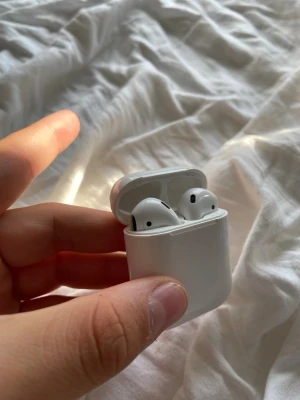 Apple AirPods med laddningsetui - Säljer ett par vita Apple AirPods med tillhörande laddningsetui. Trådlösa hörlurar med stilren och kompakt design i blank plast. Perfekta för musik, poddar och samtal på språng. Laddningsetuiet är smidigt att ta med i fickan eller väskan.