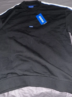 Svart sweatshirt från Hugo med blå detaljer storlek S-M - Svart sweatshirt från Hugo med blå logotyp på bröstet och blåvita ränder längs axlarna. Tröjan har rund halsringning och ribbade muddar vid ärmslut och nederkant. Perfekt för en stilren och sportig look. Aldrig använd, 