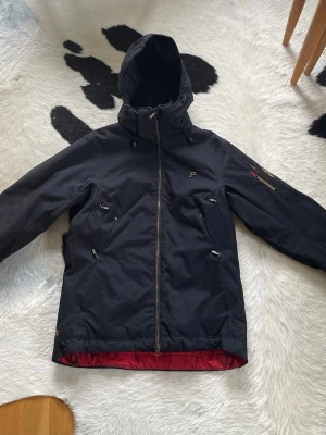 Svart vindjacka från Peak Performance i M - Snygg svart vindjacka från Peak Performance med huva och dragkedja framtill. Storlek M. Jackan har flera fickor med dragkedja och röd insida. Perfekt för blåsiga dagar och outdoor-aktiviteter. Diskret logga på bröstet och ärmen. Sparsamt använd och i fint skick. 