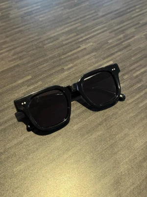 Chimi glasögon  - Klassiska svarta solglasögon från Ray-Ban med rund form och tunna metallbågar. Glasen är mörka och ger en stilren vibe, perfekt för soliga dagar. En ikonisk accessoar som alltid är trendig.