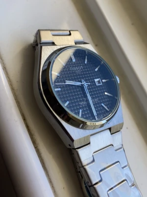 Tissot PRX automatisk klocka silver - Tissot PRX automatisk klocka med borstat silverfärgat armband i metall och svart urtavla med rutigt mönster. Klockan har datumfönster och stilren design, perfekt för dig som gillar klassiska accessoarer.