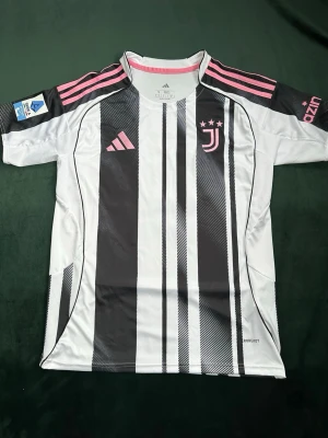 Juventus matchtröja Adidas kenan Yildiz - Juventus matchtröja från Adidas i svartvitt med rosa detaljer och klassiska ränder. Tröjan har korta ärmar, rund hals och klubbmärke samt Adidas-logga på bröstet. Tillverkad i lätt och ventilerande funktionsmaterial, perfekt för fotbollsträning.