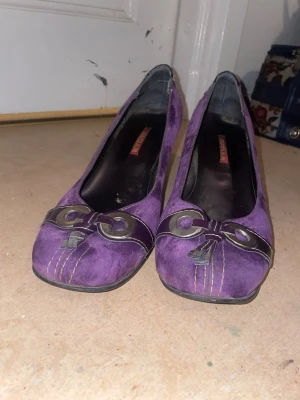Lila mockapumps med dekor - Snygga lila pumps i mocka från Sedar med dekorativa detaljer framtill i form av metallringar och sömmar. Skorna har en klassisk klack och rundad tå, vilket ger en retroinspirerad look. Perfekta för dig som vill sticka ut med färg och stil.