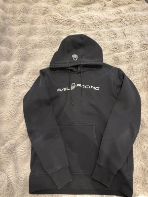 Svart Sail Racing hoodie med tryck - Svart hoodie från Sail Racing med vit logga och text på bröstet. Klassisk känguruficka framtill, dragsko i huvan och ribbade muddar. Perfekt för en avslappnad och sportig stil. Passar även för S. Vilket jag har på alla mina kläder.