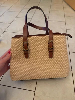 Beige handväska med bruna detaljer - Snygg beige handväska i flätat material med tre fack. Helt oanvänd!! Ett längre band medföljer💞💞