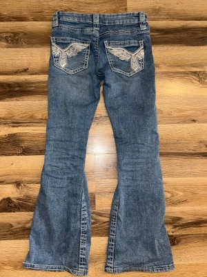 Blå bootcut jeans från Gina Tricot - Säljer ett par blå bootcut jeans från Gina Tricot med snygga broderade detaljer på bakfickorna. Jeansen har klassisk femficksmodell, dragkedja och knapp med blommönster. Tillverkade i slitstark denim med vingar på bakfickor 