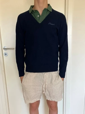 Mörkblå Peak Performance tröja - Mörkblå stickad tröja från Peak Performance med v-ringning och broderad logga på bröstet. Tröjan har långa ärmar och ribbade muddar. Perfekt att slänga på över en skjorta eller t-shirt för en clean look. Storlek L men är som en barn storlek 170. 100% lammull.