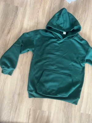 Mörkgrön hoodie med magficka - Snygg mörkgrön hoodie med klassisk huva och stor magficka framtill. Hoodien har ribbade muddar vid ärmslut och nederkant. Perfekt för en avslappnad och stilren look.