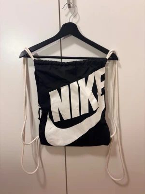 Svart gympåse från Nike - Snygg svart gympåse från Nike med stort vitt logotryck och klassisk swoosh på framsidan. Väskan har vita snören och en dragkedjeficka på sidan som har stort ficka. Perfekt för träning eller skola, lätt att bära på ryggen och rymmer det viktigaste.