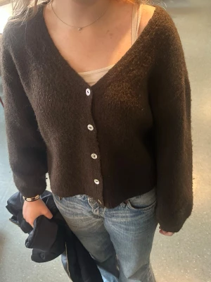 Brun kofta med knappar från Gina tricot - Mysig brun kofta med v-ringning och vita knappar framtill. Koftan har en fluffig textur, långa ärmar och en avslappnad passform. Perfekt att slänga över ett linne eller t-shirt för en chill look. Passar även M