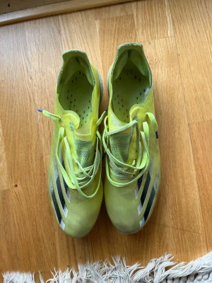 Gula Adidas fotbollsskor med snörning - Säljer ett par gula Adidas fotbollsskor med klassiska svarta och vita ränder på sidan. Skorna har snörning och ovandelen är i ett lätt, syntetiskt material som ger bra bollkontroll. Perfekta för dig som vill sticka ut på planen!