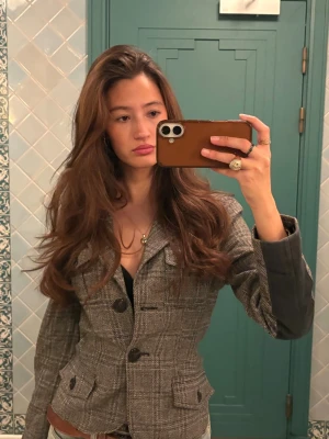 Grå rutig kavaj med knappar - Snygg grå kavaj med rutigt mönster och svarta knappar. Kavajen har figurnära passform, två fickor framtill och klassisk krage. Materialet ser ut att vara ullblandning och ärmarna har mörkare detaljer med lite skin på även bak. Perfekt för en trendig och stilren look.