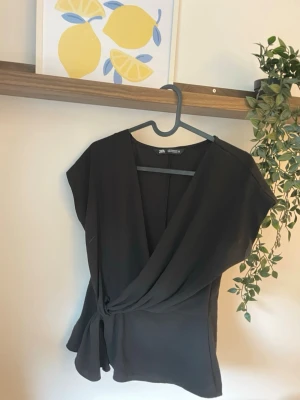 Svart omlottopp från Zara - Snygg svart omlottopp från Zara med knyt i sidan och djup v-ringning. Toppen har korta ärmar och ett stilrent, elegant fall. Perfekt att styla med både kostymbyxor och kjol för en trendig look.