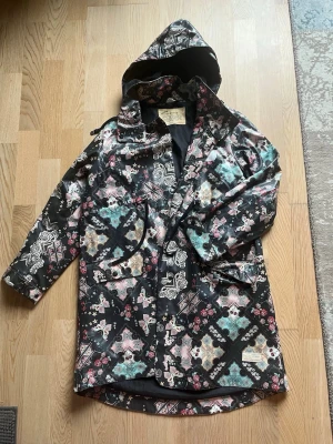 Mönstrad regnjacka från Odd Molly - Säljer en regnjacka från Odd Molly med färgstarkt mönster i svart, turkos, rosa och beige. Jackan har huva, stora fickor och knappar framtill. Materialet är vattenavvisande och jackan har en unik bohemisk design med blommor och ornament.