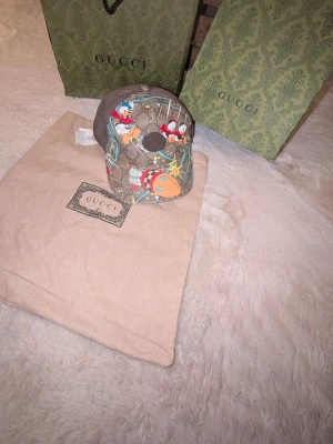Gucci x Disney keps  - Säljer en brun Gucci keps i samarbete med Disney, prydd med färgglada motiv av Kalle Anka och rymdtema. Kepsen har klassiskt GG-mönster, mesh på baksidan och justerbar rem. Perfekt för dig som vill sticka ut med en unik accessoar.