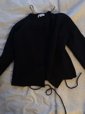 Svart omlottkofta från Gina Tricot - Mysig svart omlottkofta från Gina Tricot med långa ärmar och knytband i midjan. Koftan har en enkel och stilren design, perfekt att slänga över en topp. Materialet ser ut att vara mjukt och lite fluffigt.