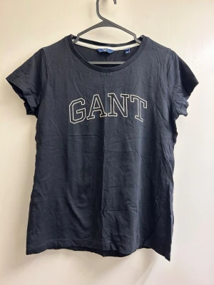 Svart GANT t-shirt med tryck - Svart t-shirt från GANT med klassiskt tryck framtill. T-shirten har rund halsringning och korta ärmar. Tillverkad i mjuk bomull som känns skön mot huden. Perfekt för en avslappnad och stilren look.