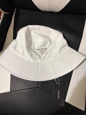 Bucket hat - Snygg vit bucket hat från Prada med klassisk logga framtill. Hatten är tillverkad i ett glansigt, vattenavvisande material och har bred skärm. Perfekt accessoar för en trendig och avslappnad stil.
