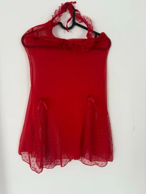 Röd mesh body med volangdetaljer - Snygg röd body i transparent mesh med volangkant runt halsen och små rosetter framtill. Axelbanden knyts i nacken och nederdelen har prickigt mönster. Perfekt för dig som vill ha något extra till underkläderna. Materialet känns lätt och luftigt.