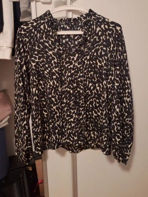 Svartvit mönstrad blus med V ringning och snören - Trendig svartvit blus med abstrakt mönster och volangdetaljer vid halsen. Blusen har långa ärmar och en lös, avslappnad passform. Perfekt för dig som gillar en sådan stil. Armarna har mudd ner till. Använd endast en gång