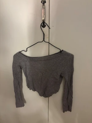 Grå ribbad croppad offshoulder topp - Säljer en grå ribbad croppad topp med offshoulder-design och långa ärmar. Perfekt för dig som gillar en trendig och enkel stil. Materialet är mjukt och stretchigt, vilket gör den bekväm att bära.
