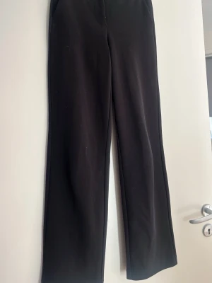 Svarta kostymbyxor från Vero Moda - Stilrena svarta kostymbyxor från Vero Moda med hög midja och raka ben. Byxorna har en diskret dragkedja fram och är tillverkade i ett mjukt, vävt material som ger en snygg siluett. Perfekta för en clean och modern look.