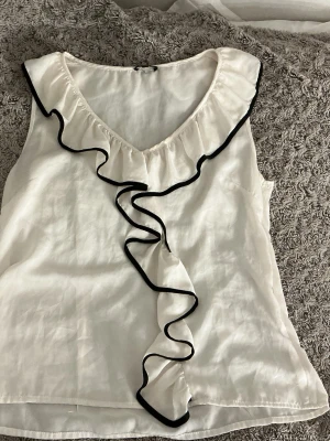 Vit volangblus från Stockh lm - Säljer en vit ärmlös blus från Stockh lm med snygga volangdetaljer längs v-ringningen och framtill, kantade med svart. Perfekt för dig som gillar en feminin och chic stil. Blusen har en mjuk och lätt känsla.