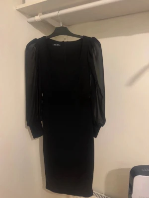 Svart klänning med puffärmar - Stilren svart klänning med transparenta puffärmar och fyrkantig urringning. Klänningen är knälång och har en figurnära passform. Ärmarna är långa och ger en elegant touch. Perfekt för dig som vill ha en klassisk men trendig look.