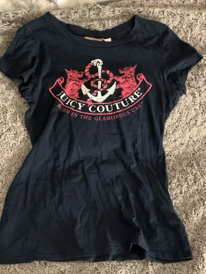 Marinblå Juicy Couture t-shirt - Snygg marinblå t-shirt från Juicy Couture med stort tryck framtill i rött och vitt, föreställande ankare och två lejon. T-shirten har korta ärmar och rund hals. 