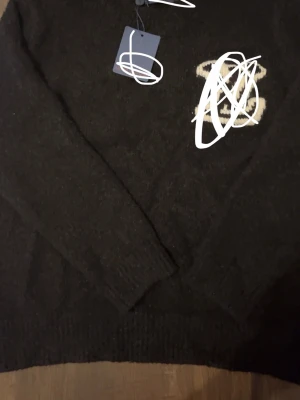 Svart stickad tröja - Svart stickad tröja med lång ärm och ribbade muddar. Tröjan har en broderad detalj i vitt och beige på bröstet samt en diskret logga. Perfekt för dig som gillar exklusiva märken och klassisk stil.