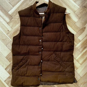 Brun manchester dunväst från Stenströms - Snygg brun dunväst från Stenströms i mjuk manchester. Västen har ståkrage, knäppning med svarta knappar och två snedställda fickor framtill. Quiltad design med breda horisontella paneler ger en cool och tidlös look.