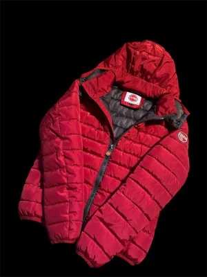 Röd pufferjacka från Colmar - Snygg röd pufferjacka från Colmar med huva och svart innerfoder. Jackan har en klassisk quiltad design, dragkedja framtill och Colmar-logga på ärmen. Perfekt för kalla vinterdagar och riktigt varm tack vare det vadderade materialet.