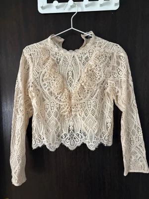 Beige spetsblus med volangdetaljer zara - Supersnygg beige spetsblus med lång ärm och hög hals. Blusen har vackra volangdetaljer framtill och ett unikt mönster i spetsen. Perfekt för dig som gillar romantisk och bohemisk stil. Materialet är genombruten spets i polyester.