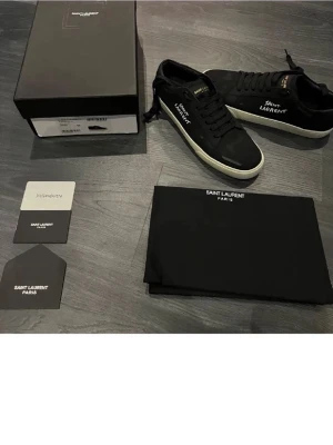 Svarta Saint Laurent sneakers - Snygga svarta sneakers från Saint Laurent med vit sula och logga på sidan. Klassisk låg modell med svarta skosnören och detaljer i vitt. Kommer med originalkartong och dustbag. Perfekta för dig som gillar stilrena och exklusiva sneakers.