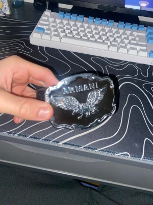 Oldschool Svart Armani bälte med stor örnspänne - Snyggt svart bälte från Armani med ett stort, blankt spänne i silverfärg och en detaljerad örnlogga. Bältet har en stilren design och passar perfekt till jeans eller byxor för en trendig look.