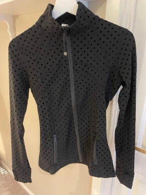 Svart prickig träningsjacka Lululemon - Snygg svart träningsjacka från Lululemon med prickigt mönster och dragkedja framtill. Tillverkad i stretchigt material med hög andel nylon och spandex för maximal komfort. Perfekt för träning eller som ett sportigt lager.