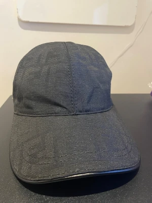 Svart keps från Fendi med logomönster - Snygg svart keps från Fendi med det klassiska FF-logomönstret invävt i tyget. Kepsen har böjd skärm och justerbar rem bak för perfekt passform. Materialet är textil med exklusiv känsla och detaljer i svart. Perfekt accessoar för en trendig streetwear-look.