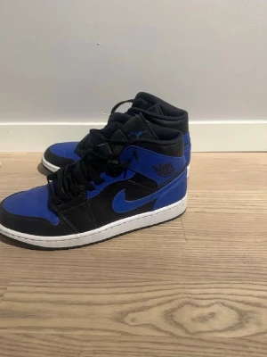 Air Jordan 1 Mid Svart/Blå - Säljer ett par Air Jordan 1 Mid sneakers i svart och blått med klassisk vit sula. Skorna har ikoniska Jordan-loggor på sidan och plösen, samt svarta skosnören. Tillverkade i en kombination av läder och syntetmaterial för en stilren look.