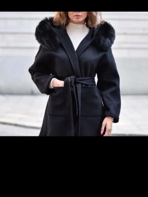 Svart kappa med fuskpäls och knytskärp - Stilren svart kappa med bred krage i fuskpäls och knytskärp i midjan. Kappan har raka ärmar och två stora fickor framtill. Perfekt för kyliga dagar och ger en lyxig känsla till din outfit.
