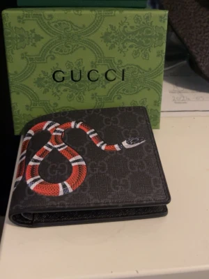 GGplånbok med ormtryck - Snygg svart plånbok från GG med klassiskt GG-mönster och en cool röd, vit och svart orm på både fram- och baksidan. Tillverkad i tåligt canvasmaterial och levereras med original grön GGi-box. Perfekt för dig som gillar statement-accessoarer.