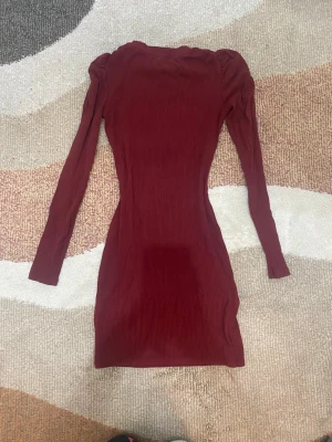 Vinröd ribbad långärmad klänning - Säljer en vinröd, ribbad kortklänning med lång ärm och puffade axlar. Klänningen har en kroppsnära passform och är tillverkad i ett stretchigt material som framhäver figuren. Perfekt för dig som gillar en enkel men snygg stil. Använd endast fåtals gånger, den är tajt men inte super.