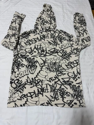 Graffiti-mönstrad hoodie från H&M - Cool hoodie från H&M med vit bas och svart graffiti-mönster över hela plagget. Hoodien har huva och långa ärmar, perfekt för dig som gillar streetstyle och unika prints. Materialet är mjukt och bekvämt, idealiskt för att chilla eller hänga med kompisar.